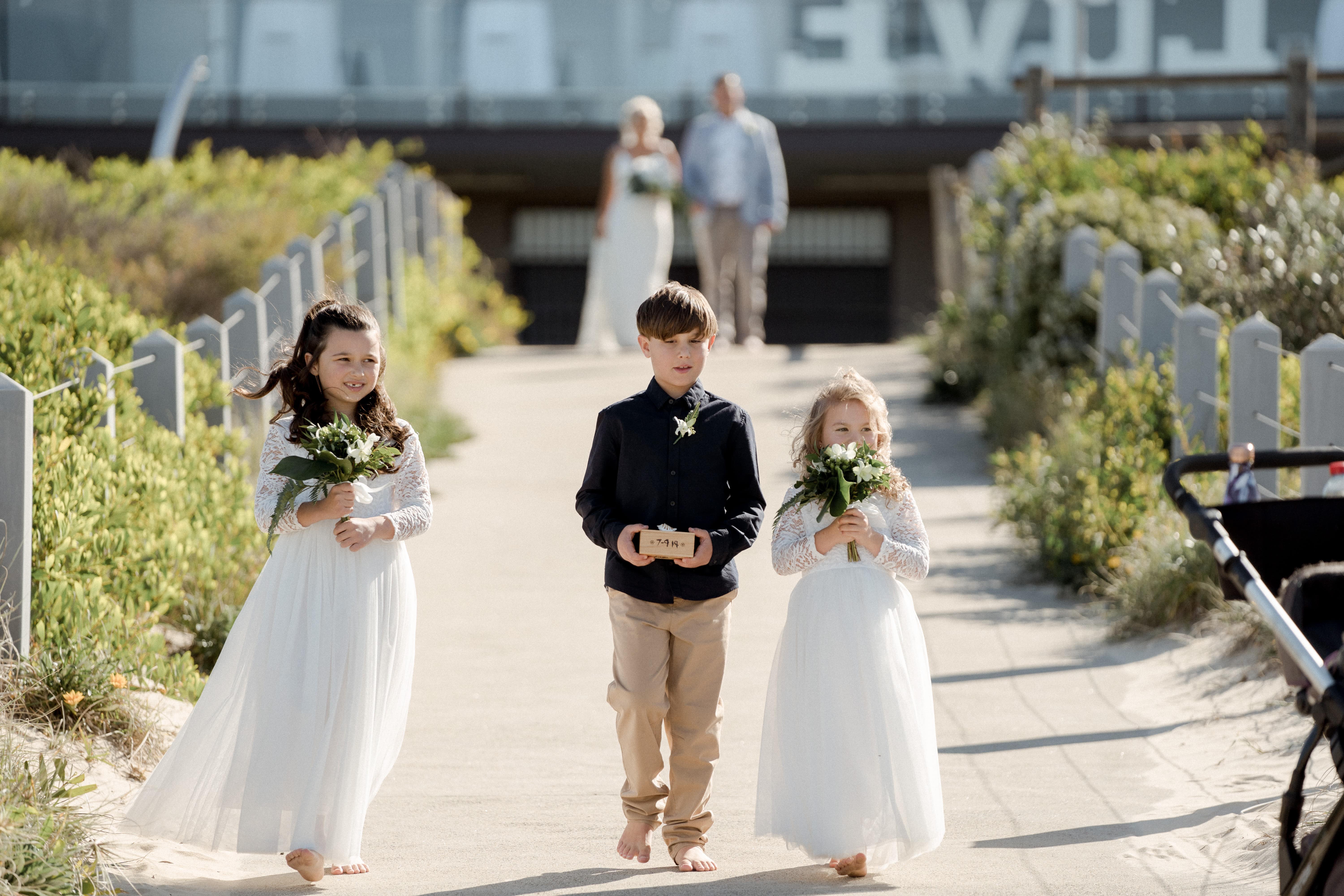 Paigeboy and flowergirls entrance before the Bride and Father of the Bride walk down the aisle to marry on the beach